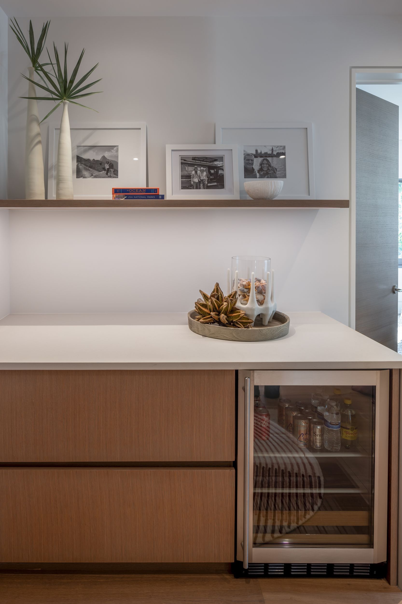 Built-in bar with light wood cabinetry, countertop, and beverage cooler. Shelf holds framed photos and vase.