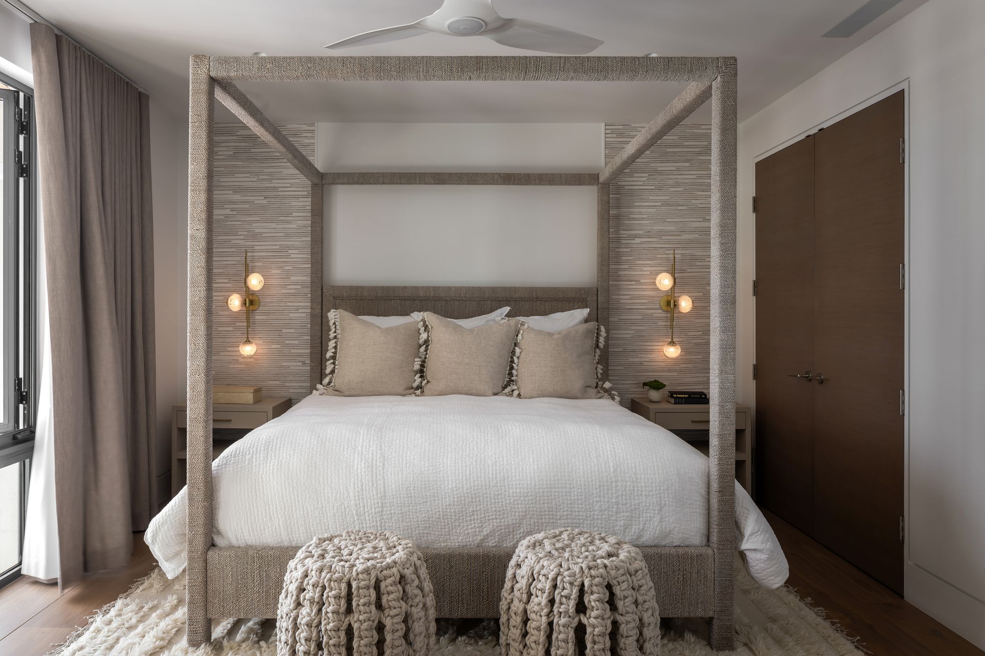 Bedroom with a canopy bed, neutral tones, wood accents, and plush bedding. Two stools sit at the foot of the bed.