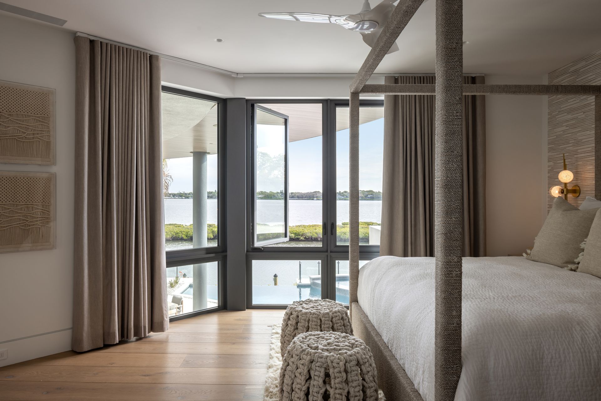 Bedroom with a four-poster bed, water view, and large windows with gray curtains. Two textured stools sit by the bed.