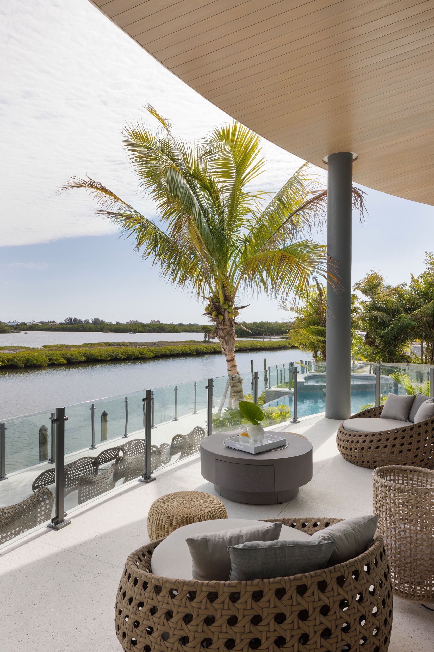 Outdoor seating area overlooking water, with wicker furniture, palm tree, and glass railing.