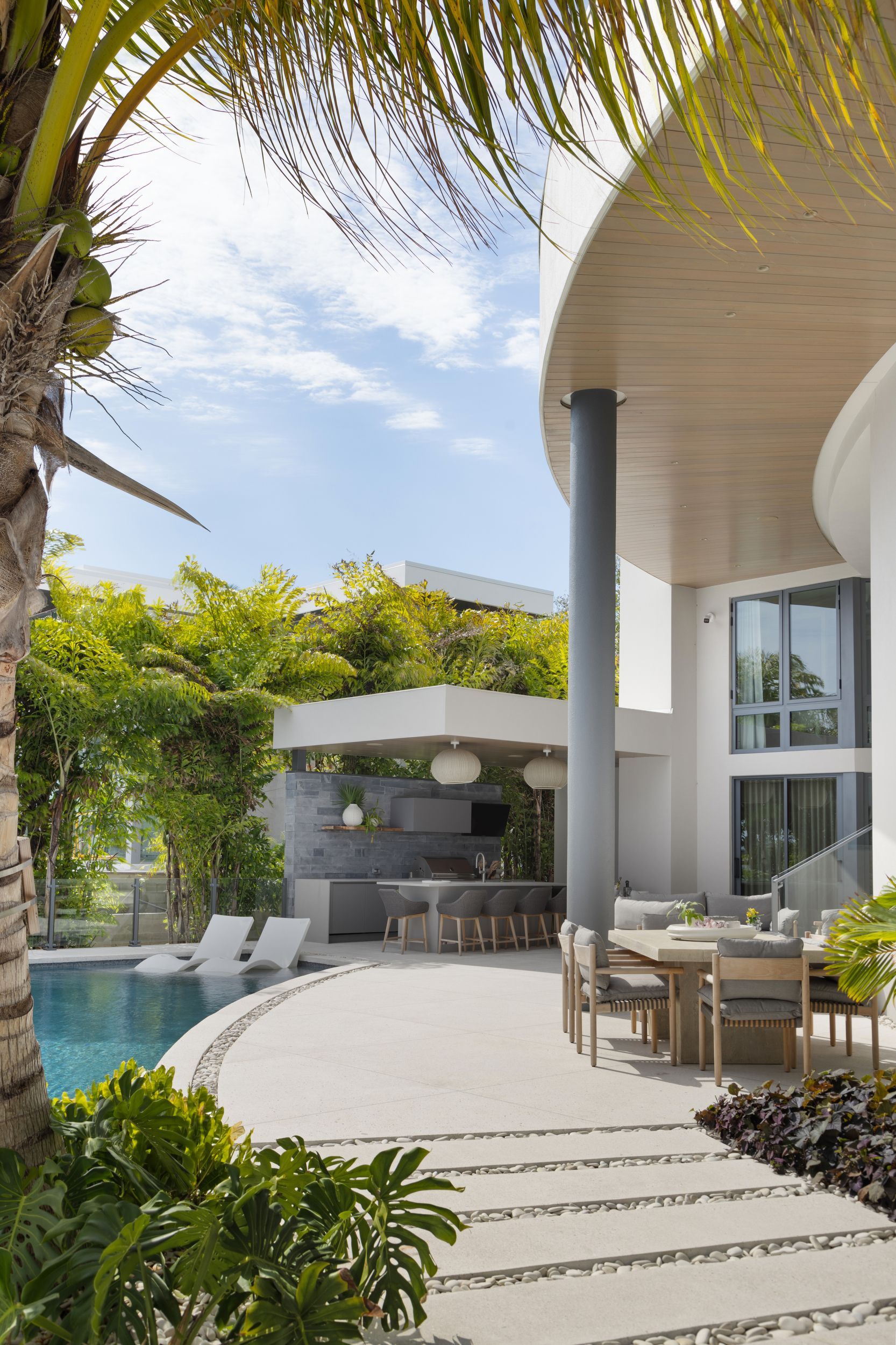 Curved patio with pool, outdoor kitchen, and dining area. Palm trees frame the sunny scene.
