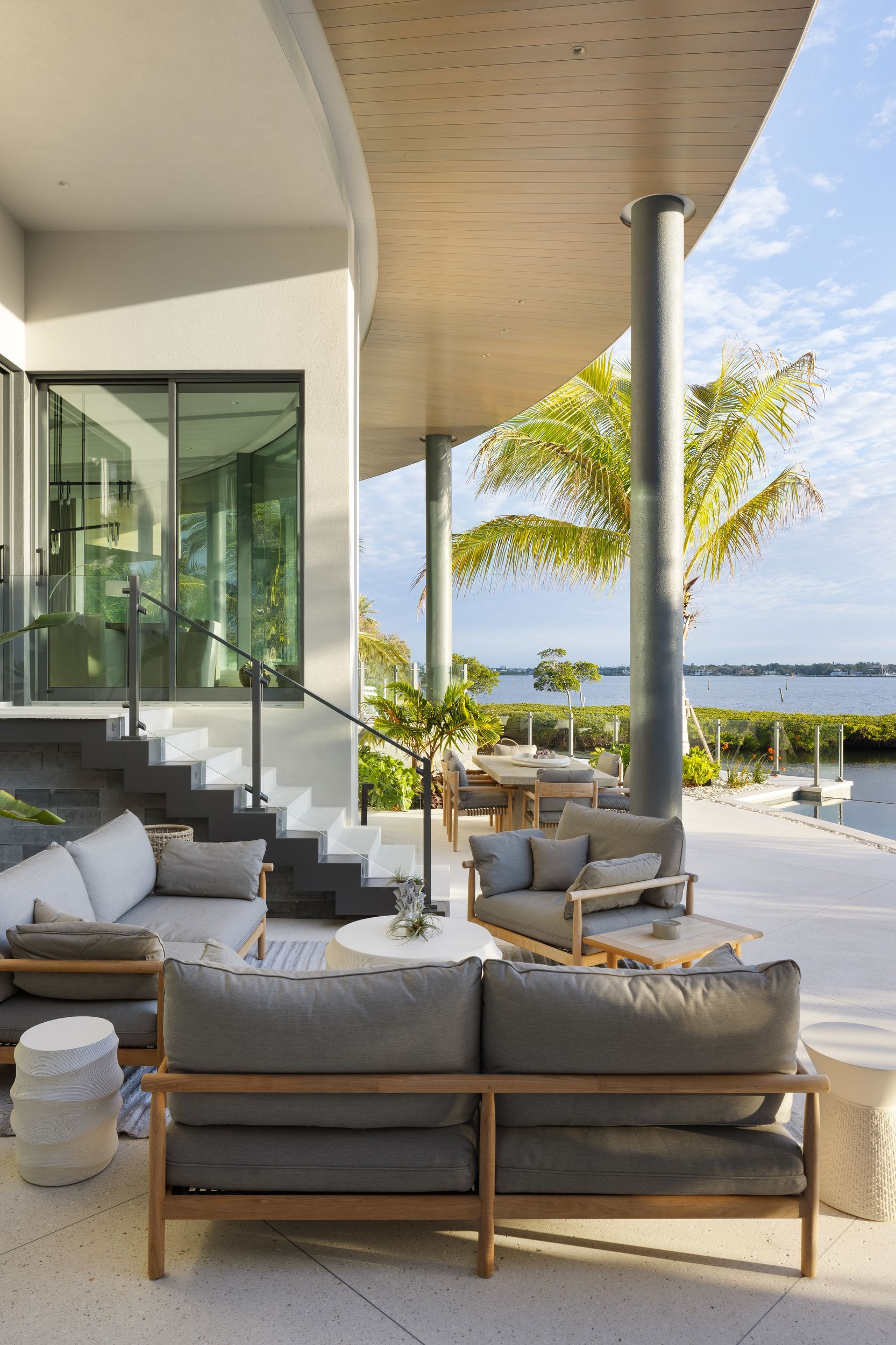 Outdoor patio with gray couches, round tables, and water view under a curved roof.