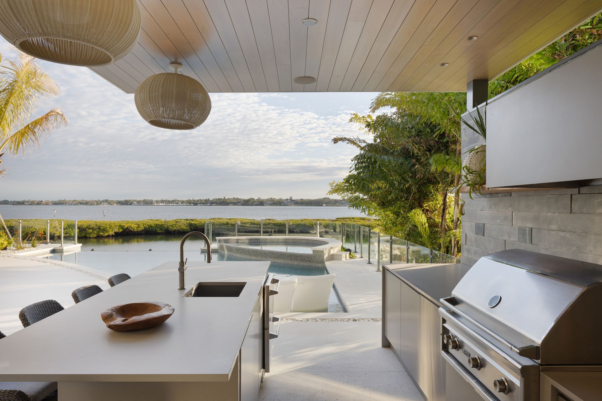 Outdoor kitchen with a view of water, pool, and plants; neutral tones, modern design.