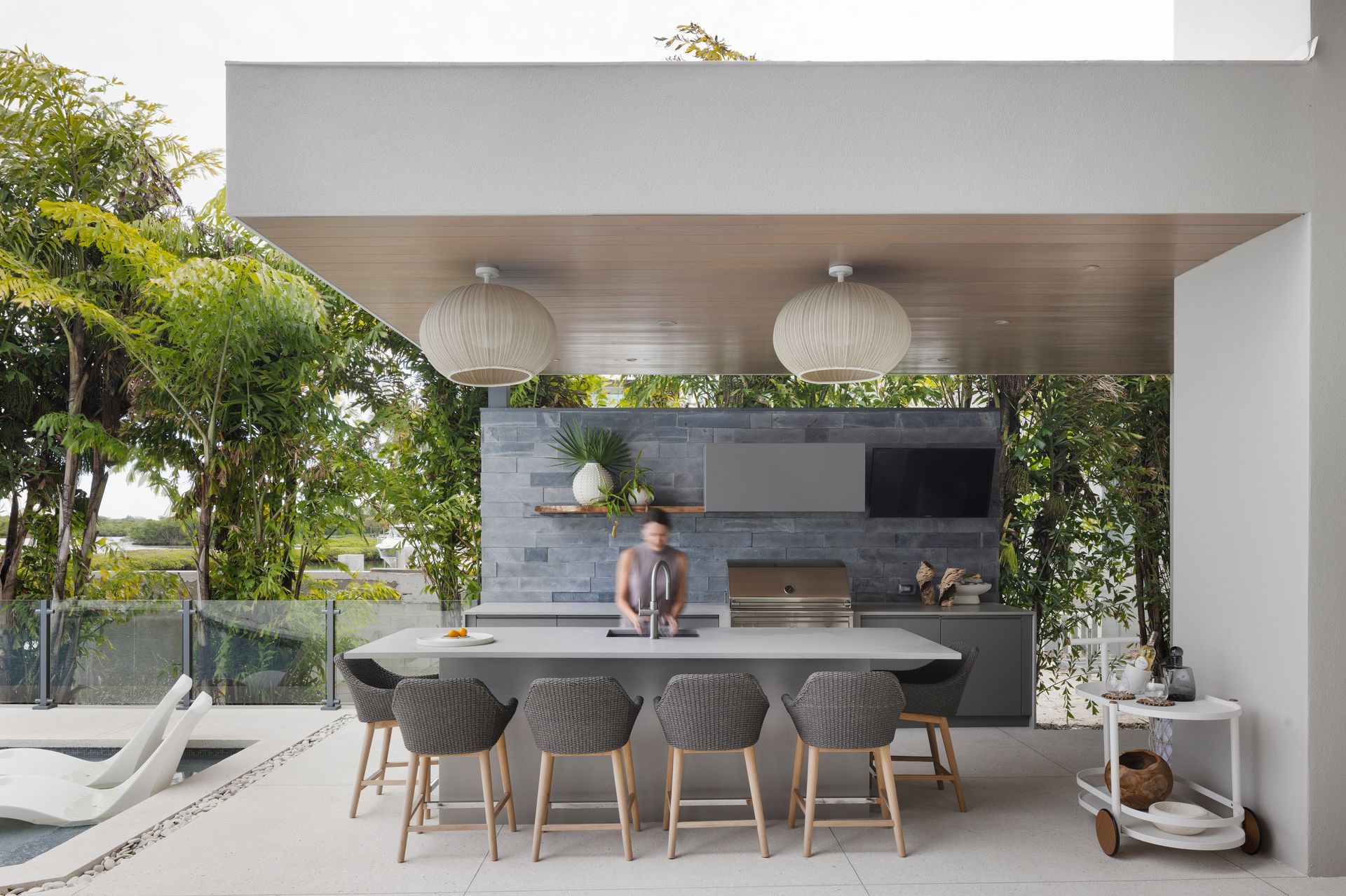Outdoor kitchen with a woman preparing food, a grill, and a bar with seating.