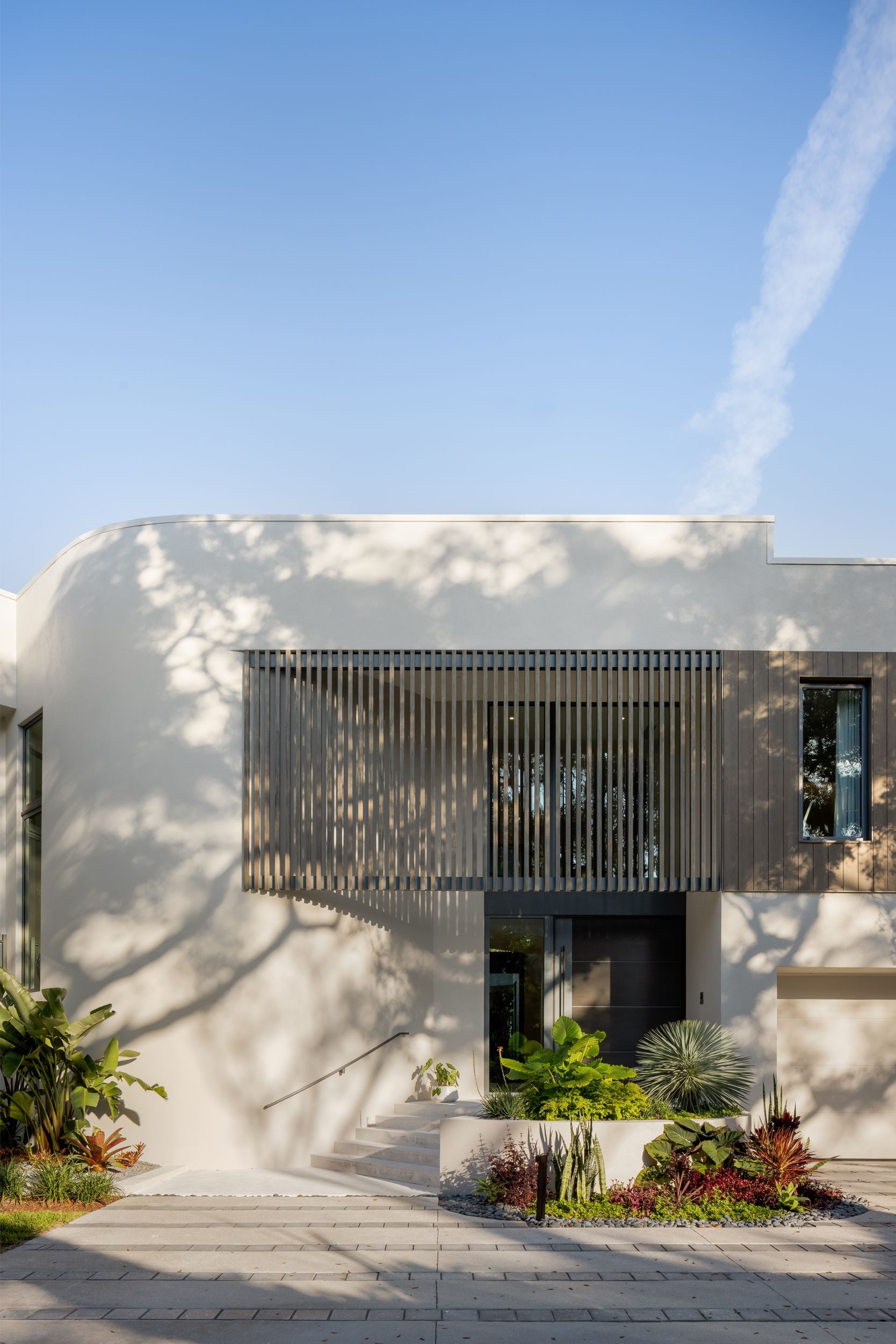 Modern white house with wooden slat facade, doorway, and foliage against a blue sky.