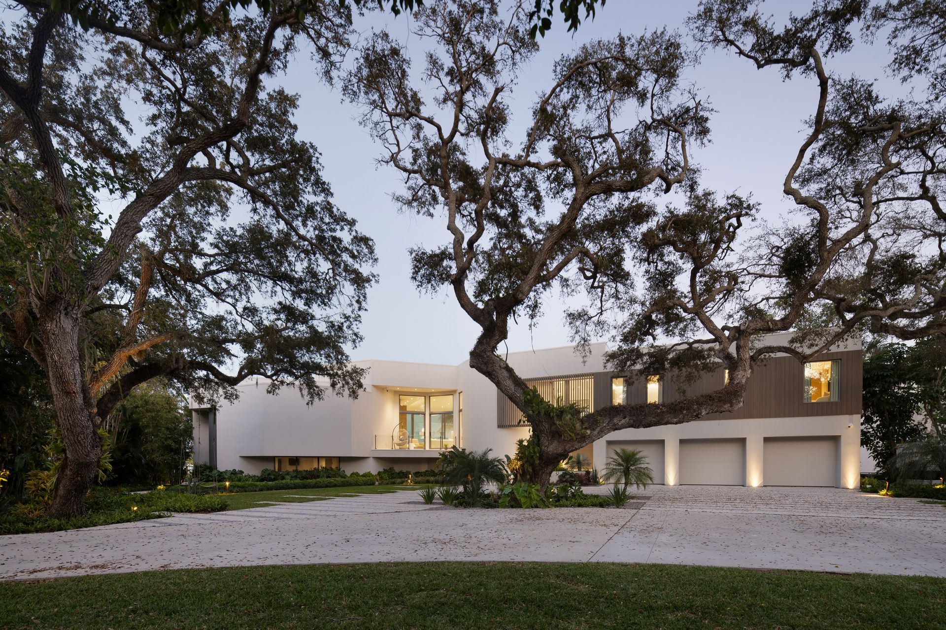 Modern white house with arched driveway, three-car garage, and large oak trees in front.