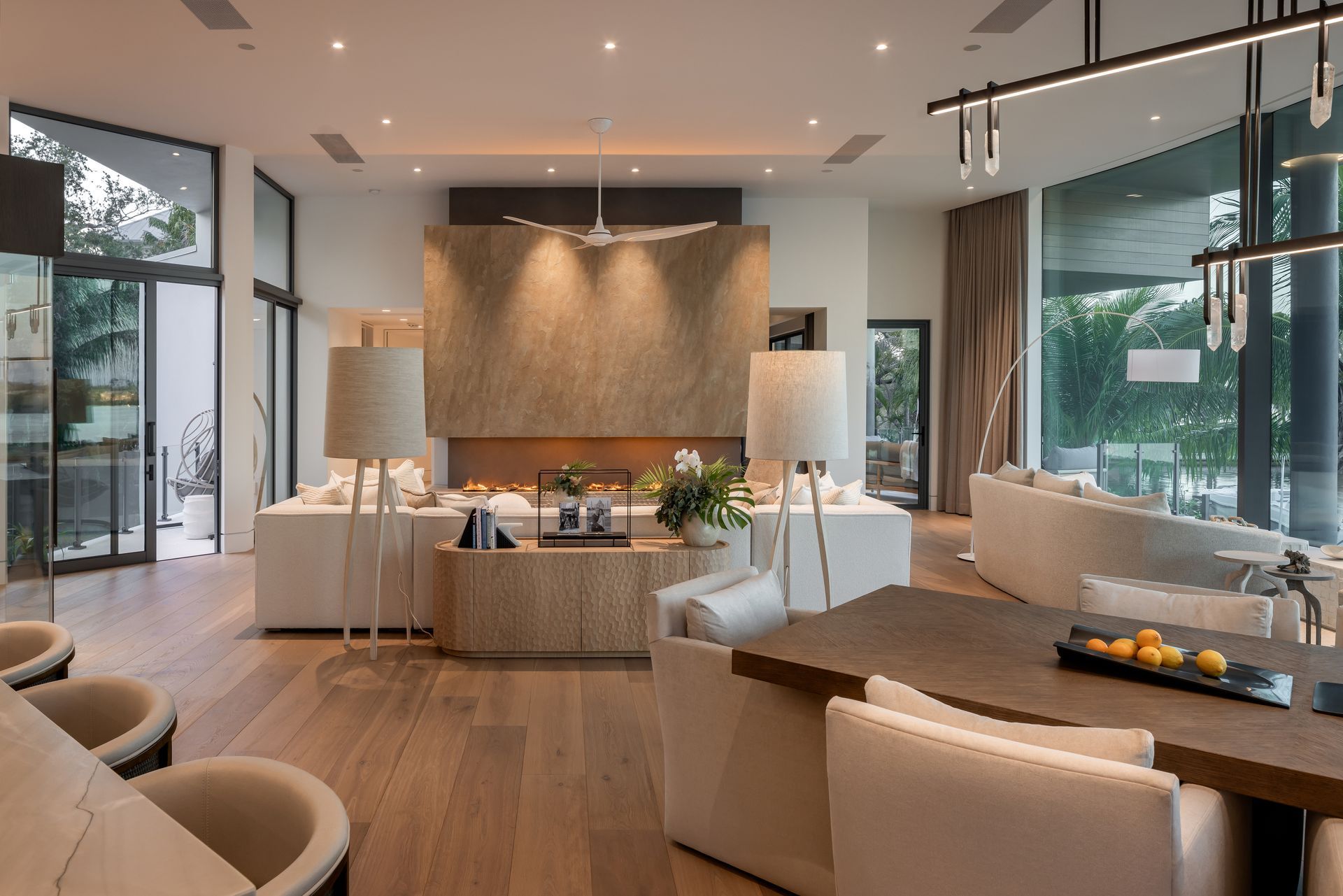 Spacious living room with a fireplace, light wood floors, and large windows. Furnished with cream sofas and a dining table.