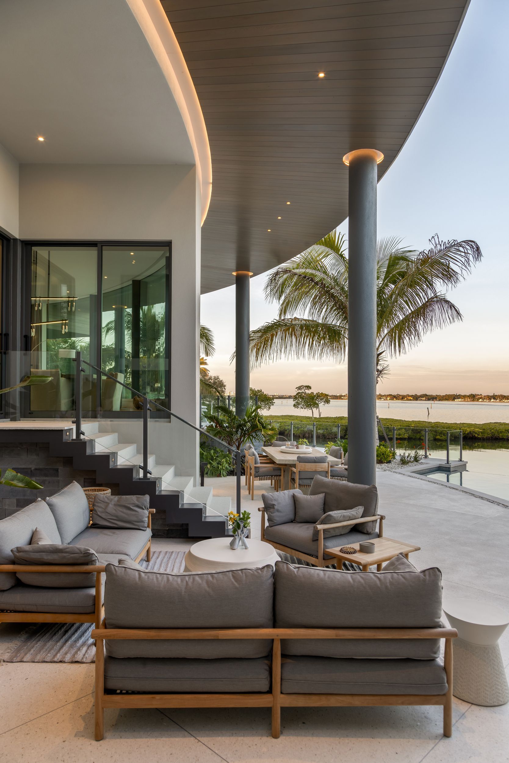Outdoor patio with gray sofas, wooden tables, and waterfront view under a curved roof.