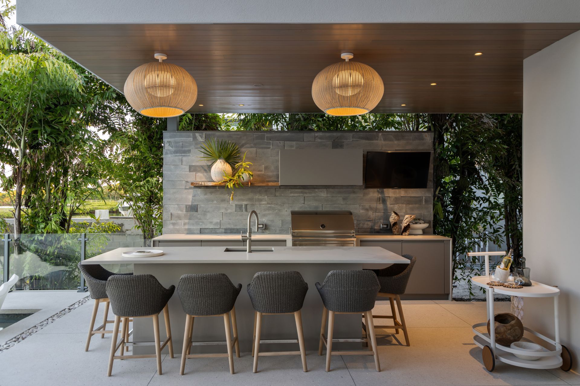 Outdoor kitchen with gray cabinets, stone backsplash, island seating, and woven pendant lights.