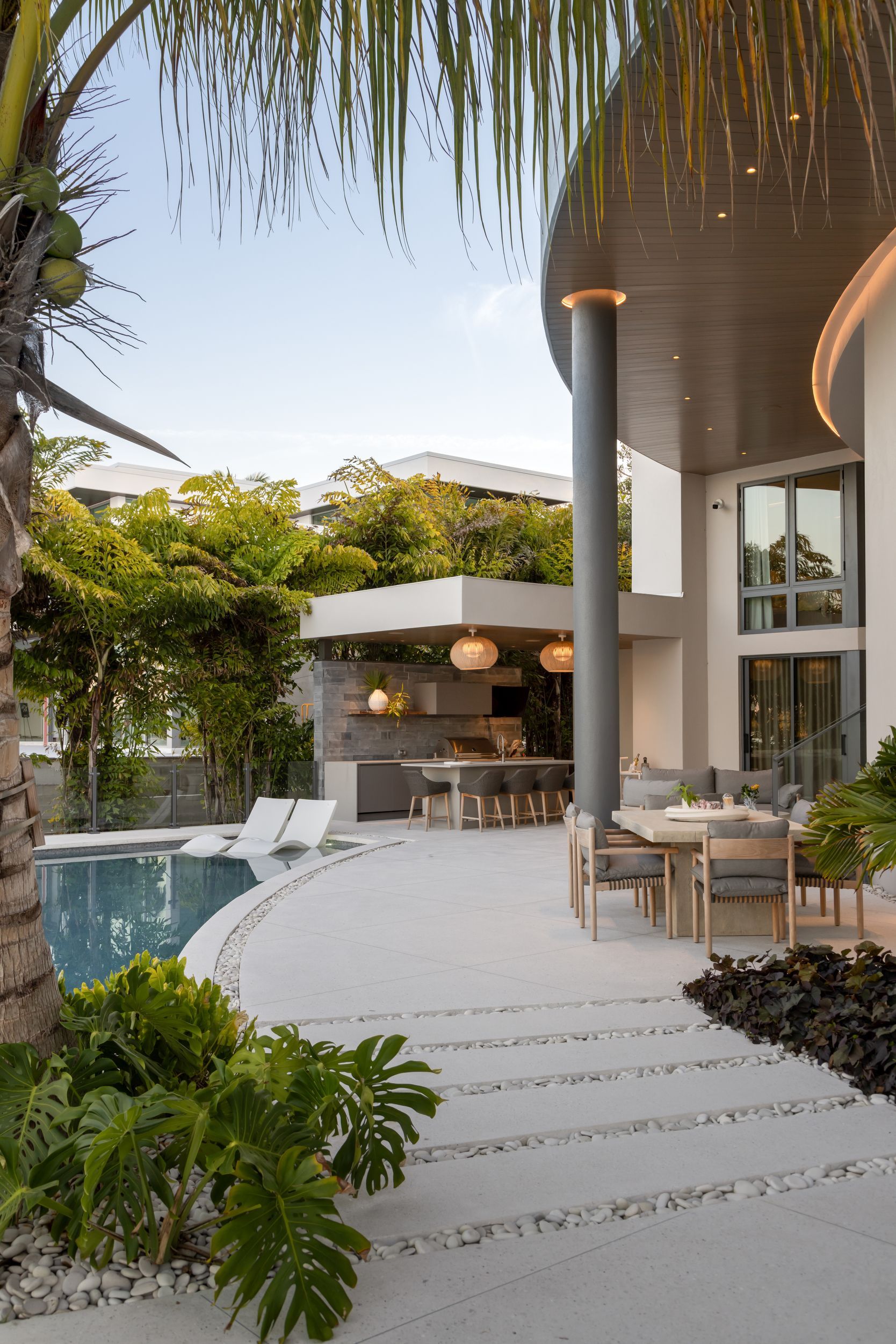 Luxury outdoor patio with pool, dining, and kitchen area. White stone path, tropical landscaping.