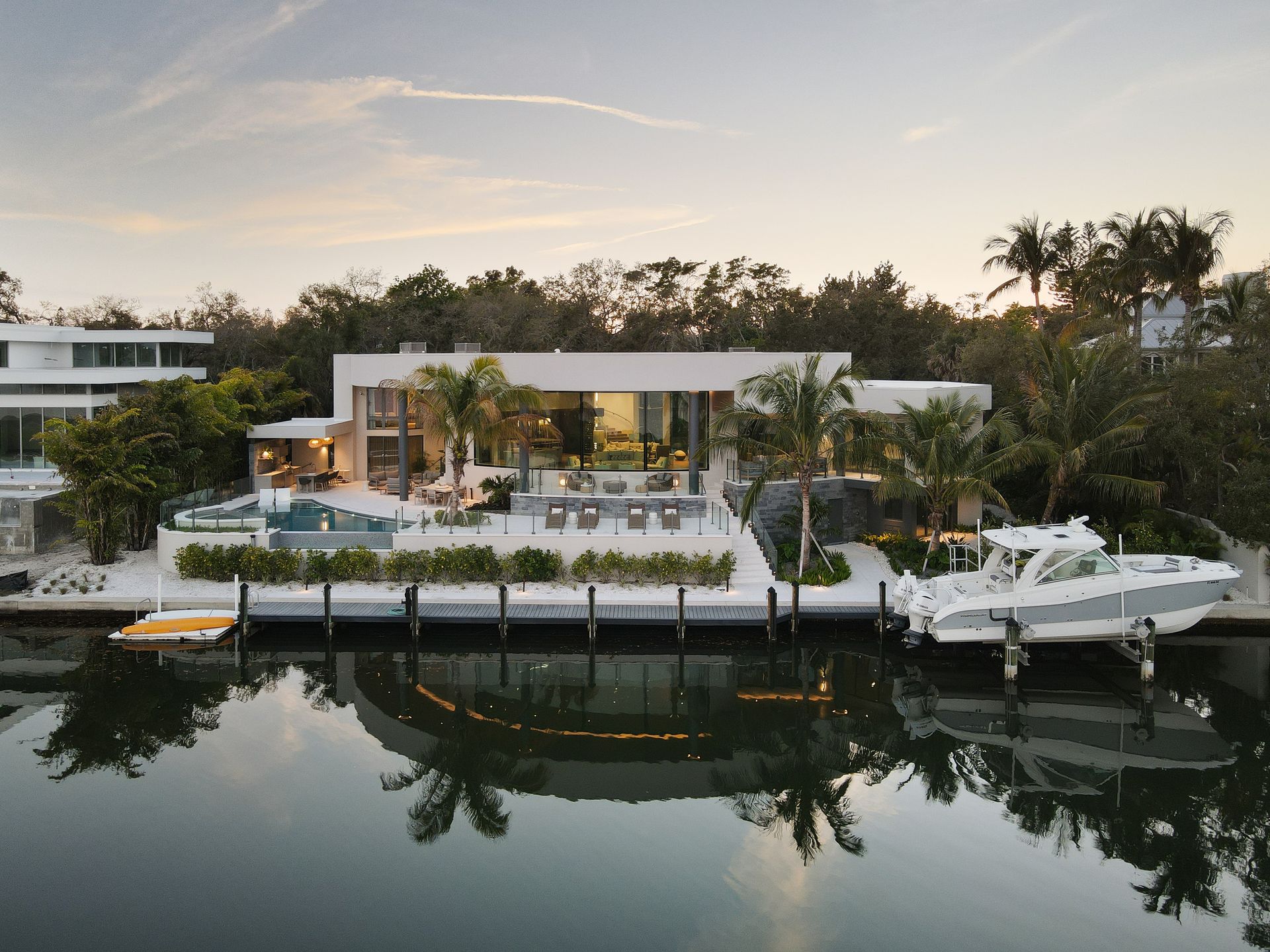 Modern waterfront home with pool, dock, and boat, reflecting in calm water.