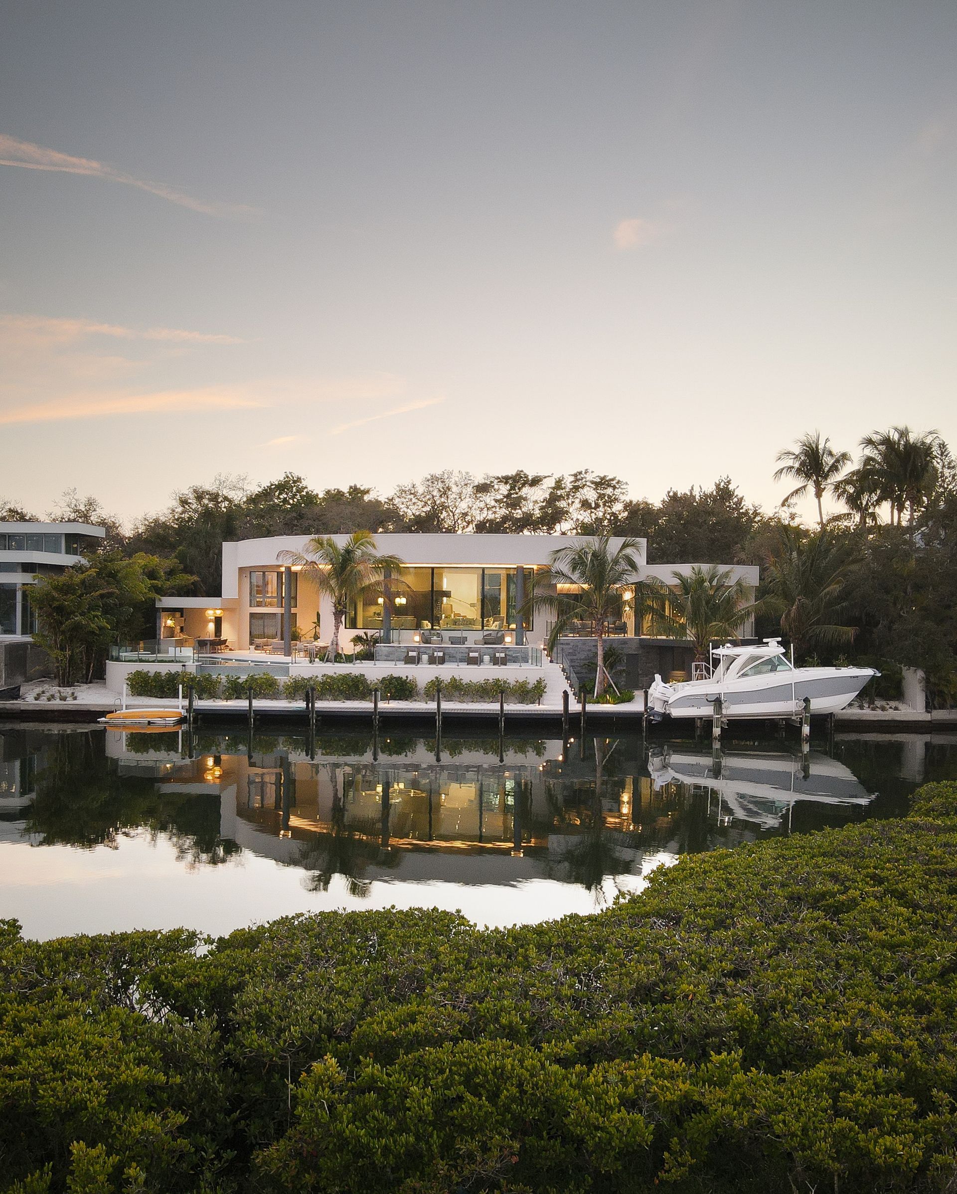 Modern white waterfront home with boat docked, surrounded by trees, reflecting in the water.
