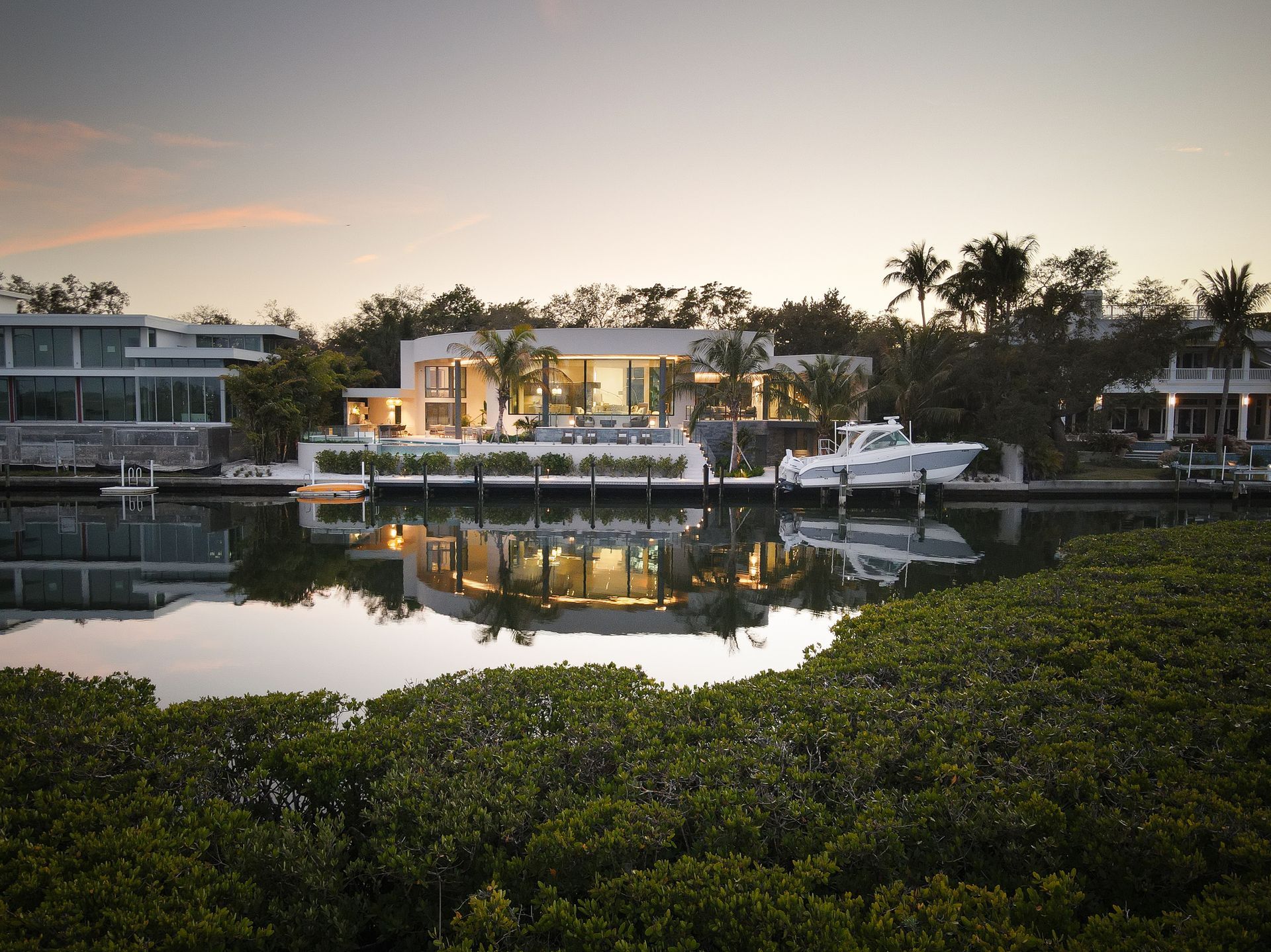 Modern waterfront home at dusk reflecting in calm water, with a boat docked nearby.