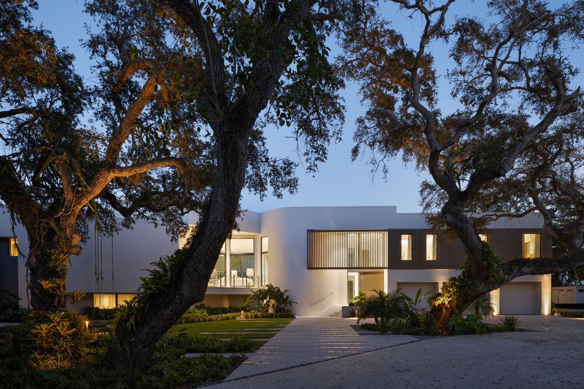 Modern white house with arched facade, illuminated at dusk, framed by large trees.