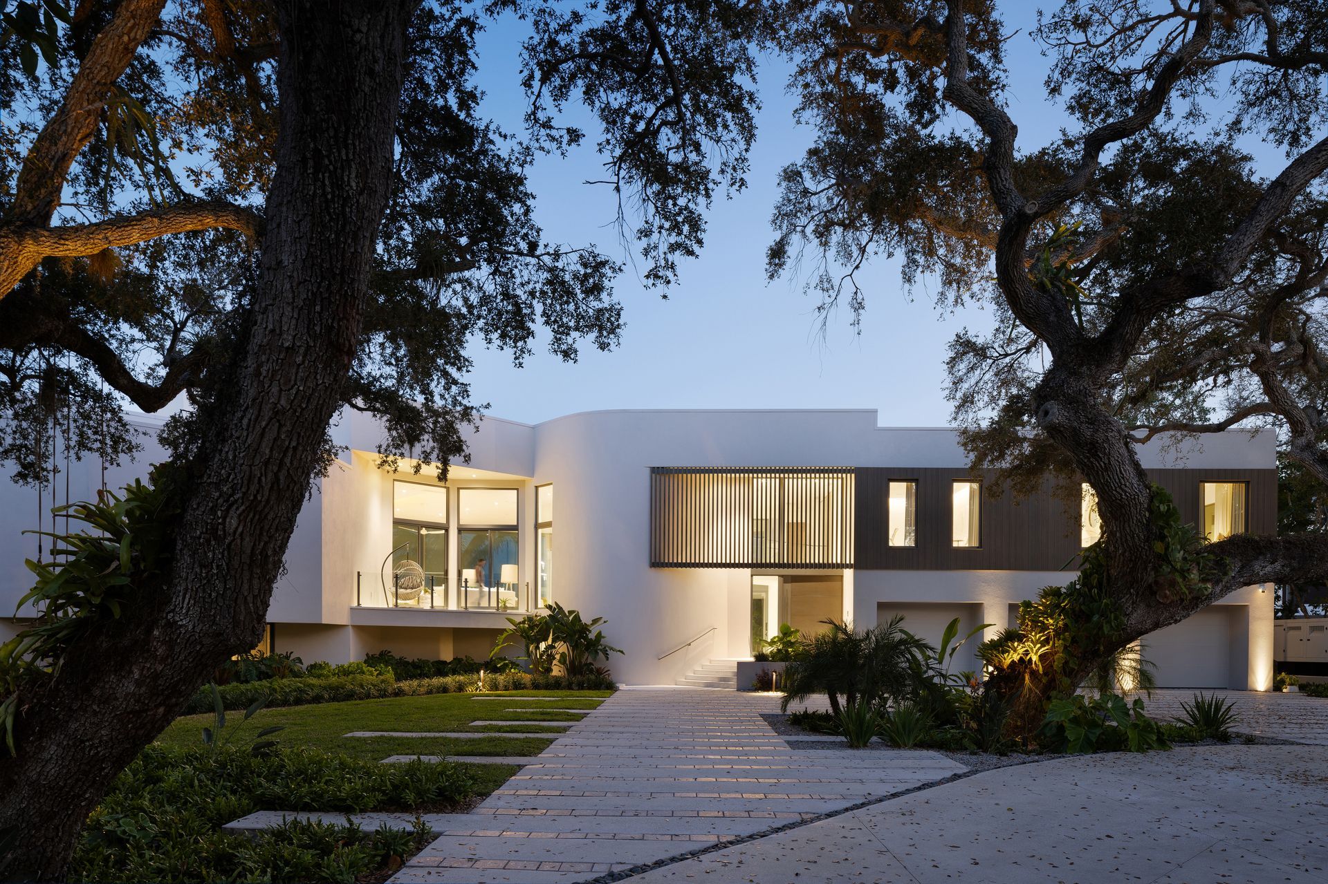Modern white house framed by large trees, lit pathway leads to entrance.