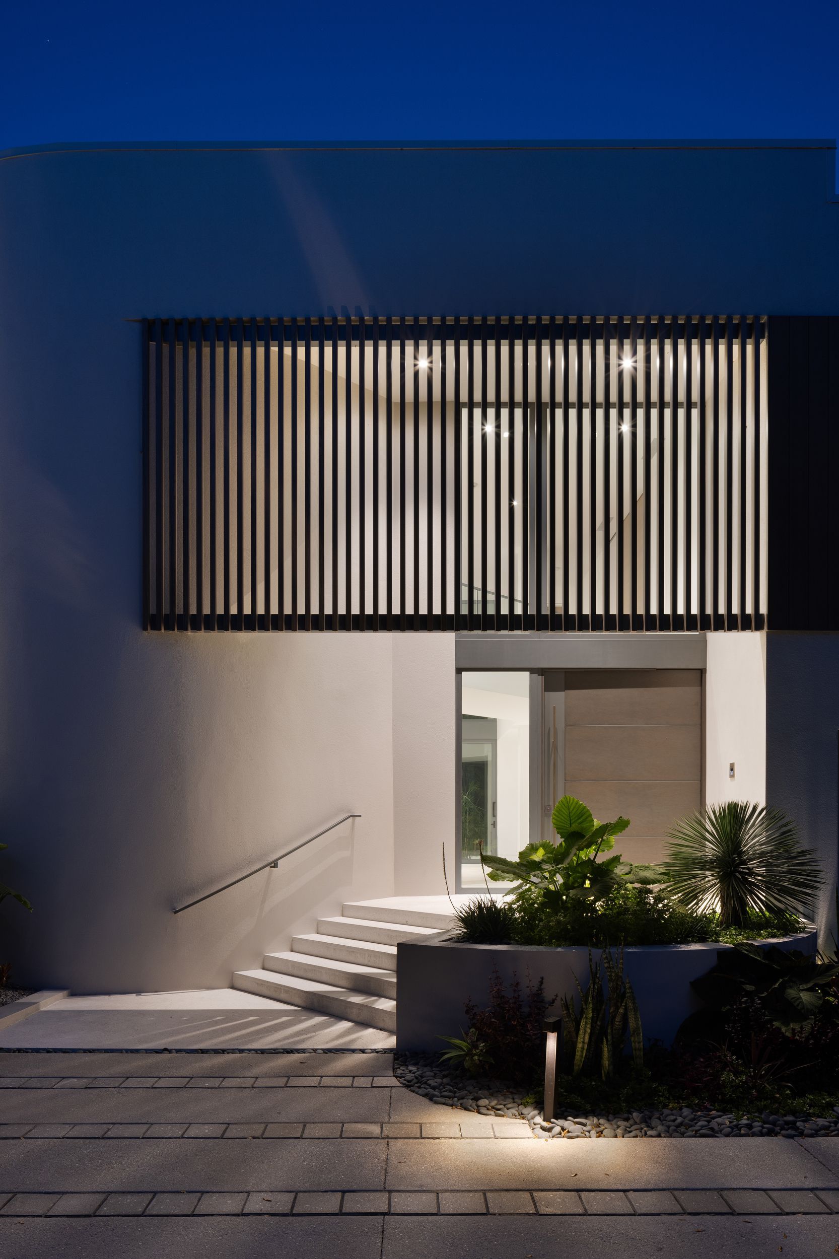 Exterior of modern white house at night, lit stairs lead to entrance with vertical slatted window above.