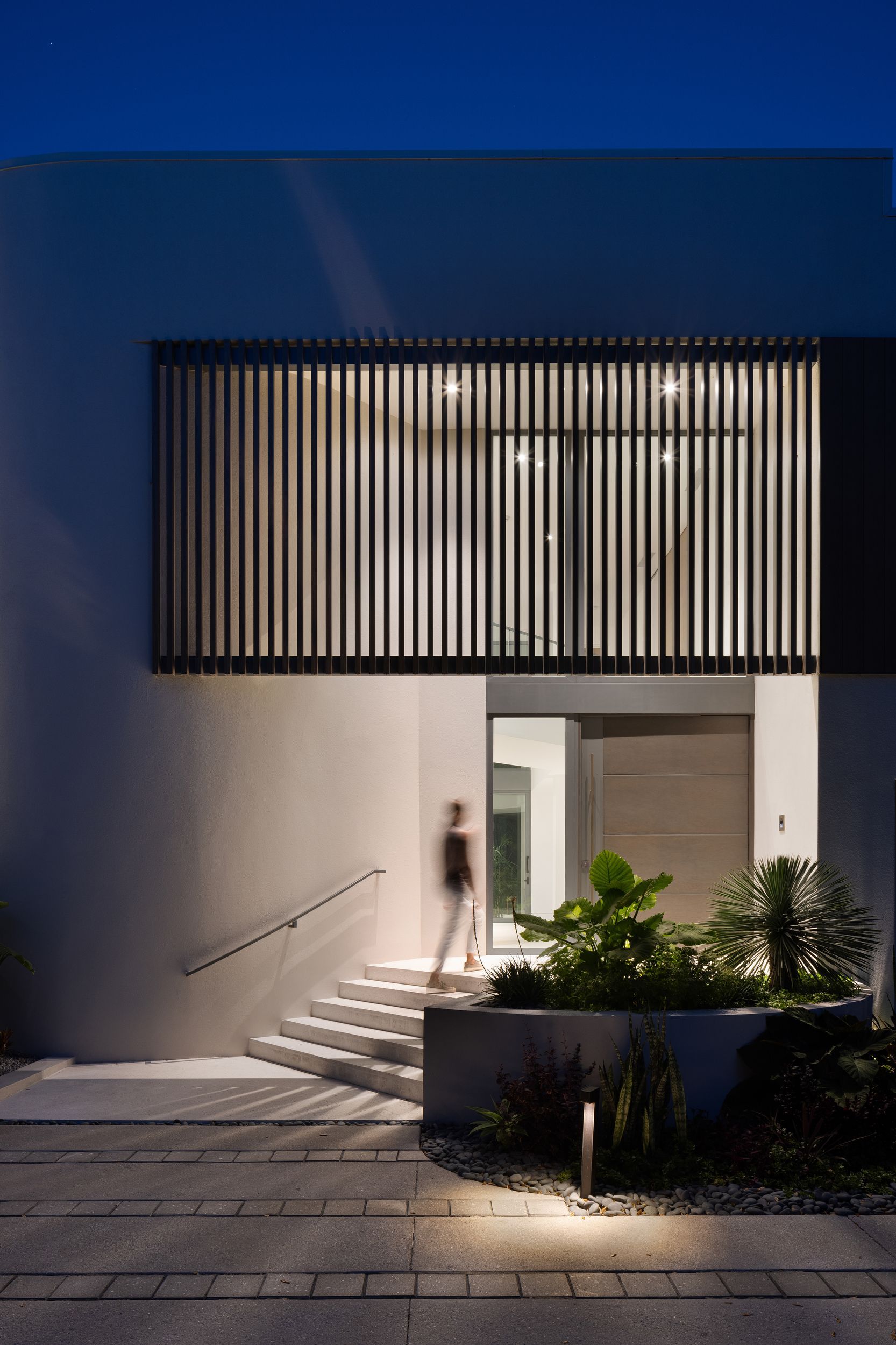 Modern home entrance at night; person entering; vertical slatted window, lit steps, landscaping.
