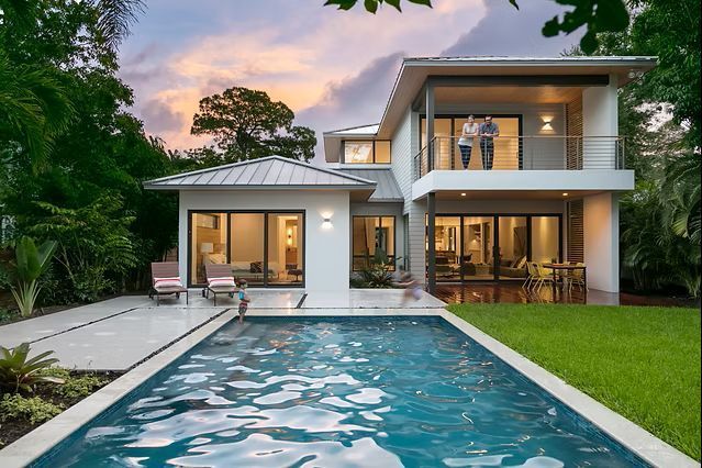 Modern white house with a pool, two-story structure, lush greenery, and a family on the balcony.