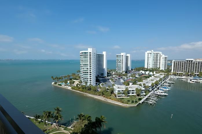 High-rise white buildings on a peninsula, surrounded by blue water. Sunny day with docked boats.