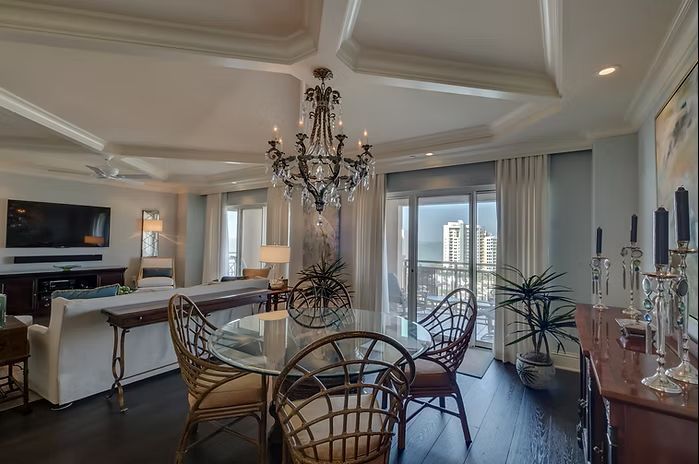 Elegant dining room with chandelier, glass table, wicker chairs, dark wood floor, and balcony view.