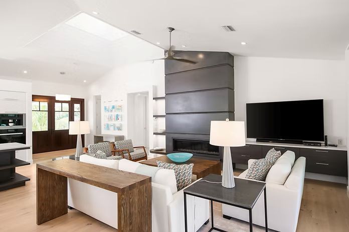 Modern living room with fireplace, TV, and white sofas. Light wood floors and large windows.