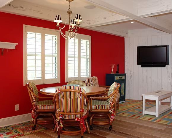 Red dining room with round table, striped chairs, and shutters. TV on white wall.