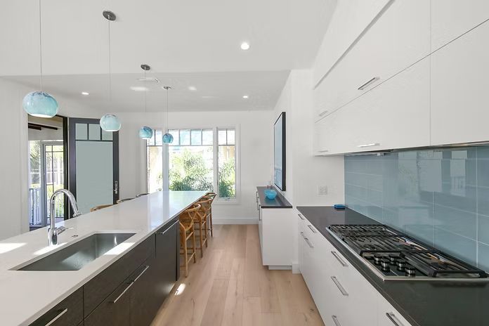 Modern kitchen with white cabinets, dark island, blue glass backsplash, and light wood flooring.
