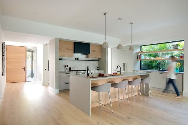 Modern kitchen with island, bar stools, light wood floors, large window, and person walking.