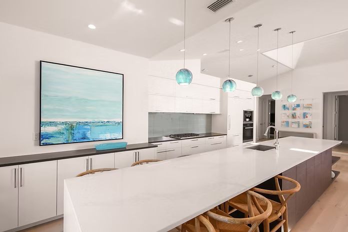 Modern kitchen with white cabinets, long island, and teal pendant lights. Artwork and bar stools complete the space.