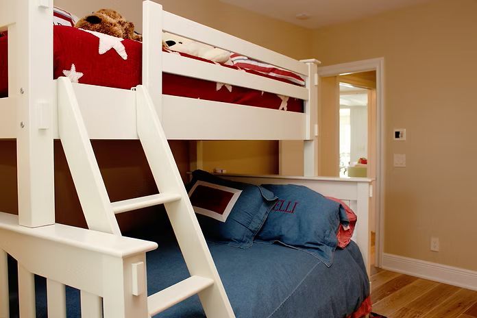 White bunk bed in a bedroom; red and blue bedding, light wood floors, beige walls.