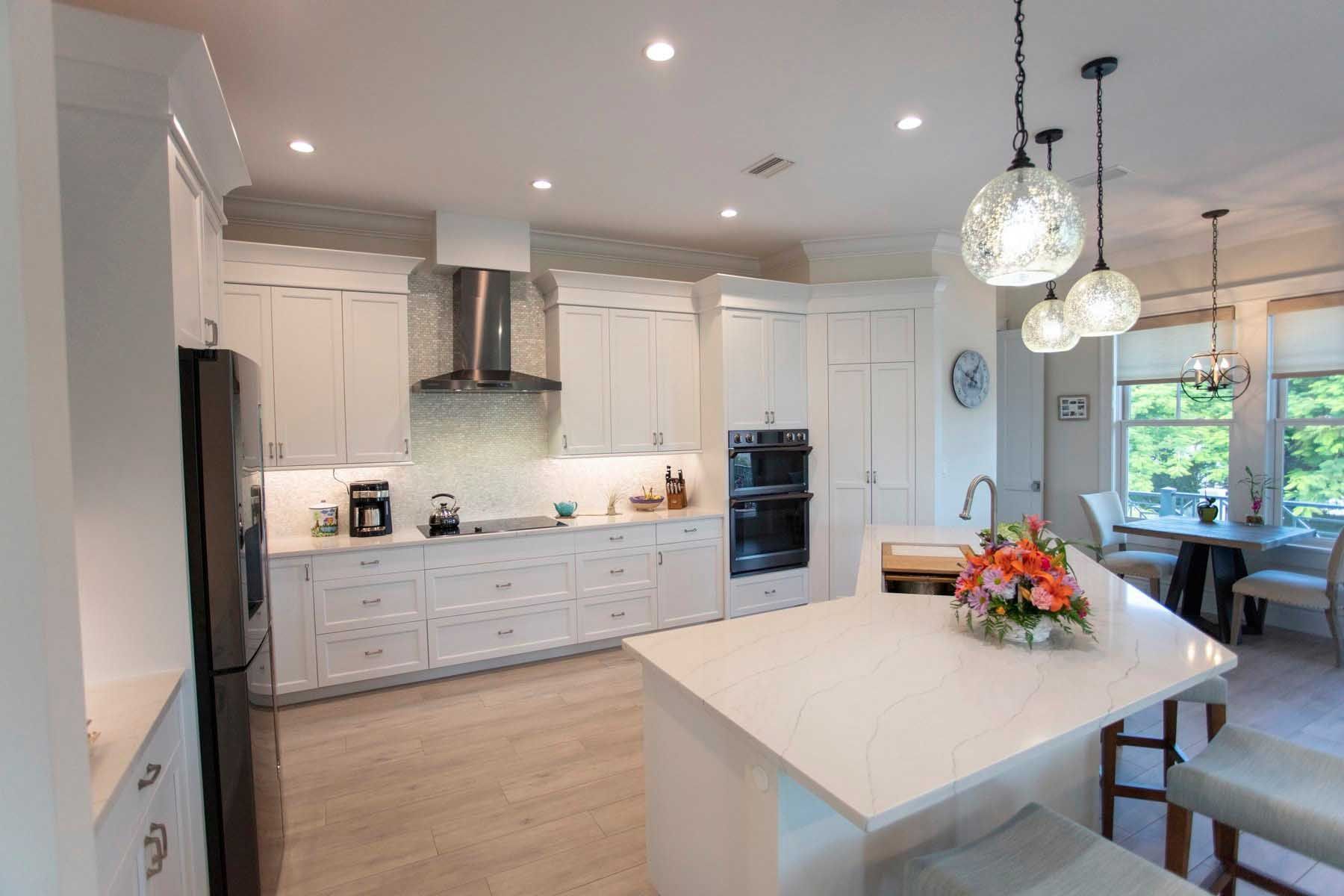 Bright white kitchen with a quartz island, cabinets, and appliances. Dining area with a table in the background.