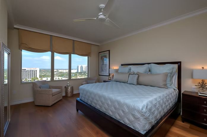 Bedroom with a bed, chair, windows, and wood flooring. Light blue bedding, beige walls, and city view.