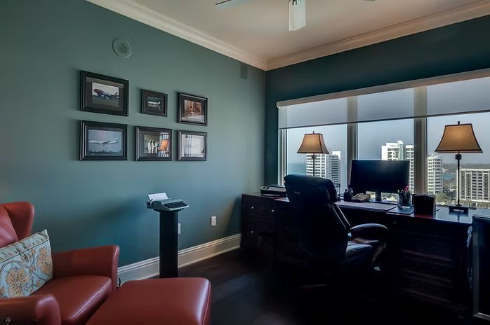 Home office with desk, chairs, artwork on teal walls, dark floors, and a city view.
