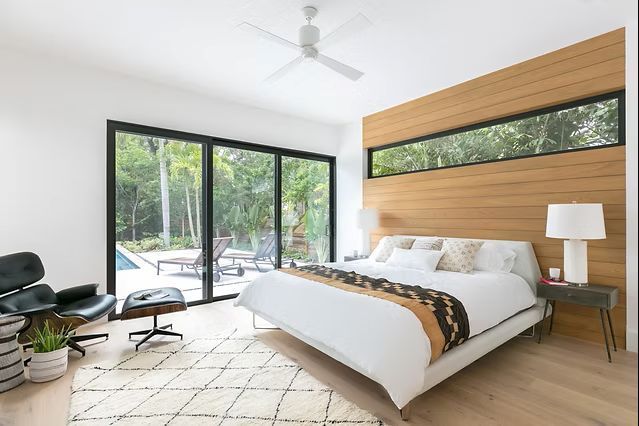 Modern bedroom with large windows overlooking a pool, wood headboard, white bedding, black chair, and rug.