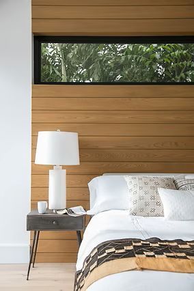 Bedroom with wood-paneled wall, small nightstand, white lamp, and bed with patterned throw. Window above.