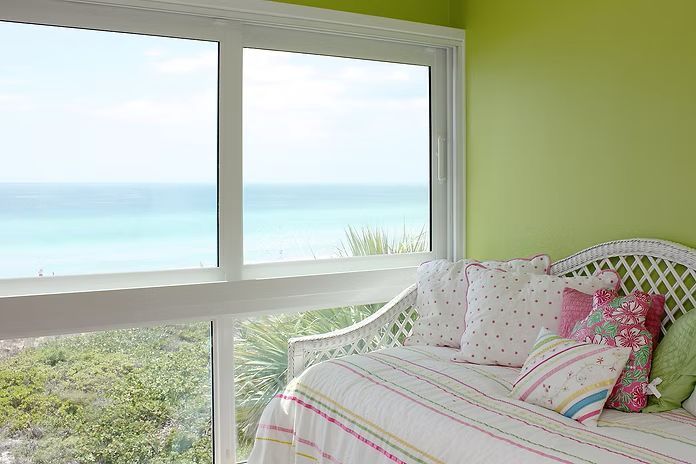 A room with a large window overlooking the ocean, green walls, and a white wicker daybed with pillows.
