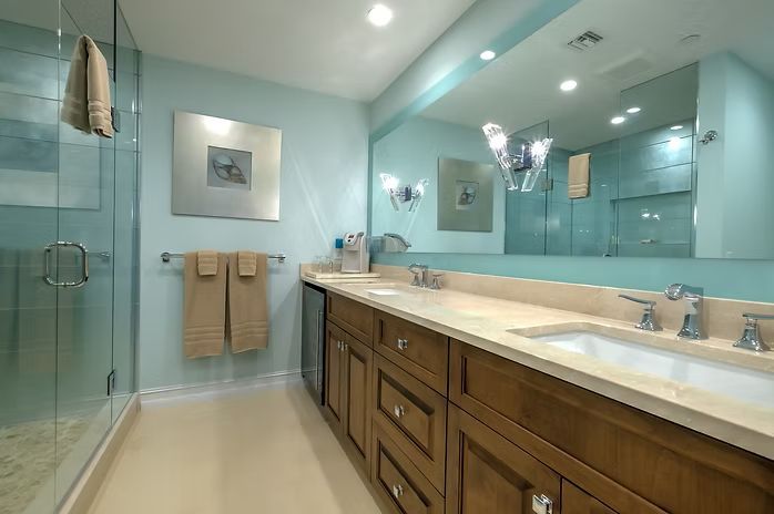 Bathroom with light blue walls, brown vanity, and large mirror. Glass shower and beige floor.