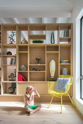 Girl using VR headset sits in front of a wooden bookcase. Yellow chair beside her, window on right.