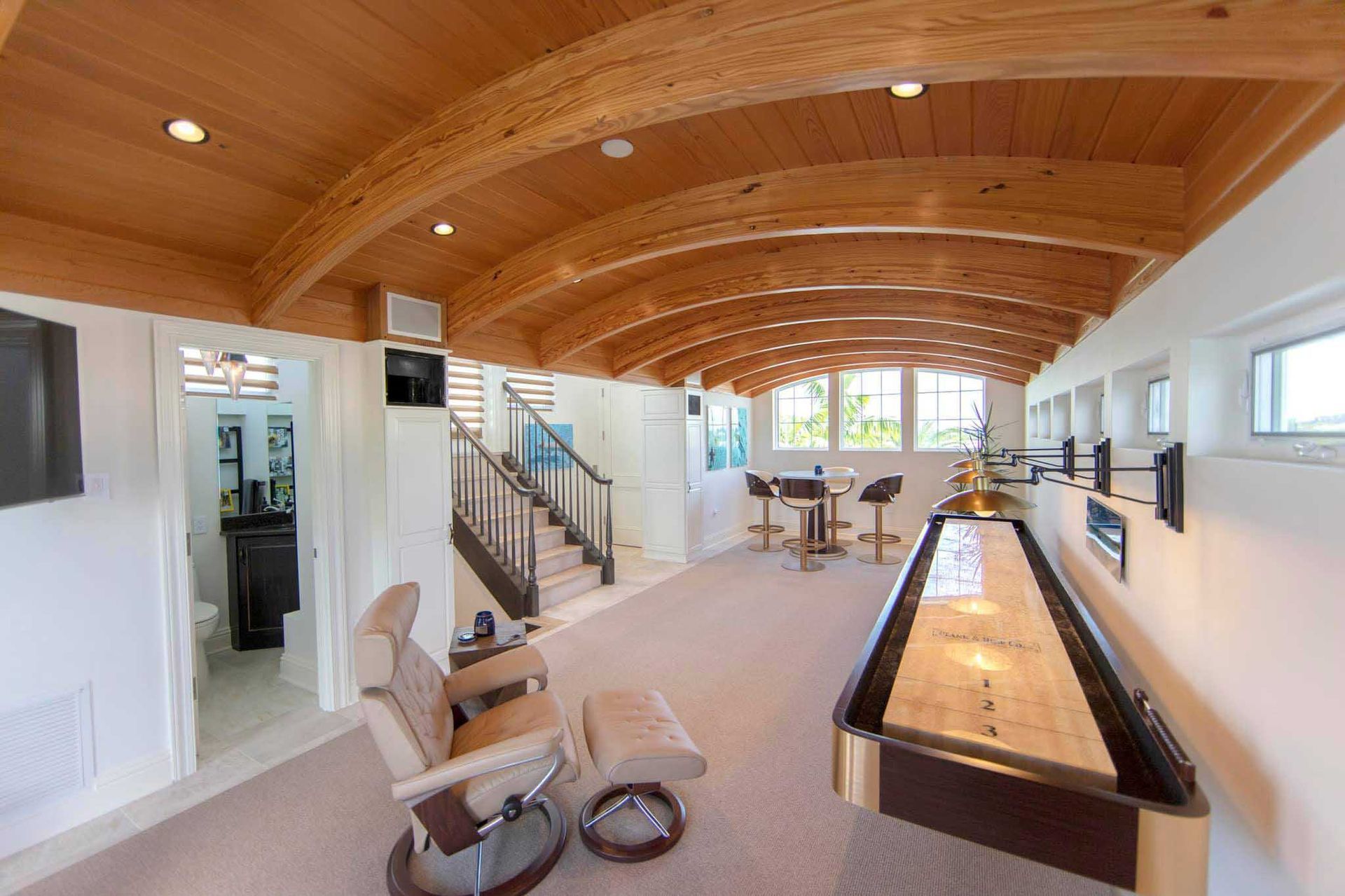 Luxurious room with shuffleboard table, lounge chair, bar, curved wood ceiling, and stairs.