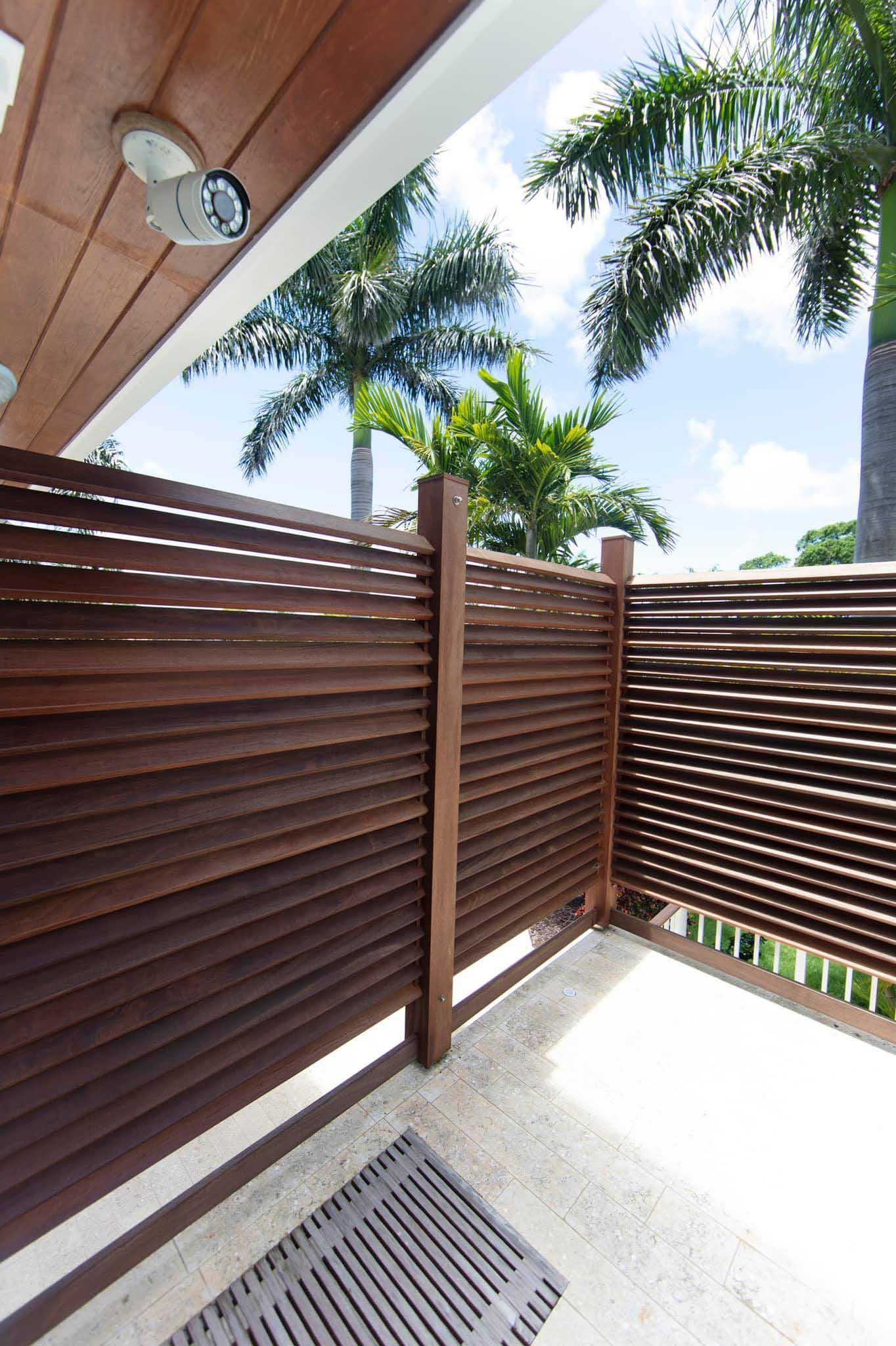 Wooden slatted fence on a patio with a palm tree backdrop under a bright blue sky.
