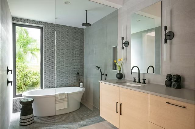 Modern bathroom with gray tiled walls, soaking tub, light wood vanity, and a large window.