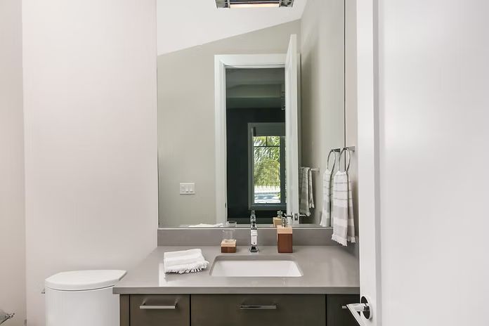 Bathroom with sink, mirror, and toilet. Gray countertop and cabinets, white walls, towels on a rack.