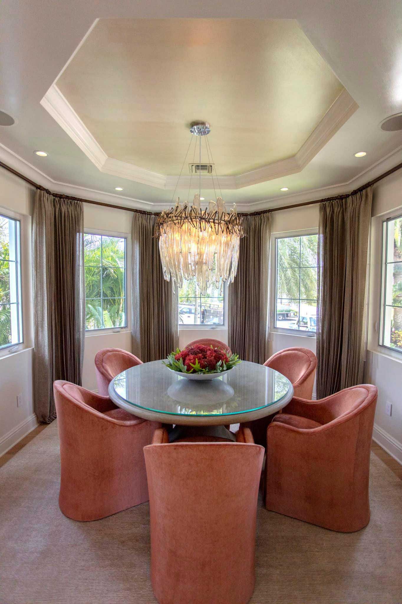 Round dining table with six coral chairs in a bright room with windows, drapes, and a chandelier.