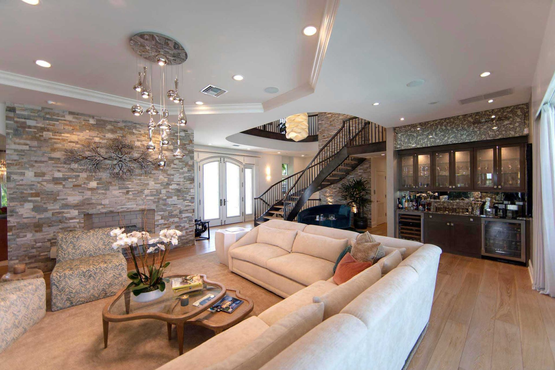 Spacious living room with stone fireplace, curved staircase, bar, and light-colored sectional sofa.