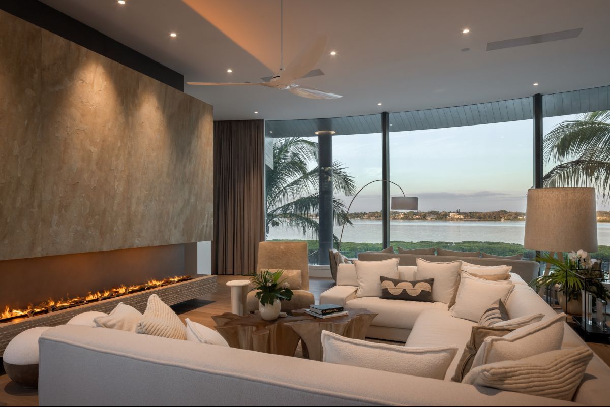 Modern living room with fireplace, curved window overlooking water, neutral tones, plush sectional sofa.