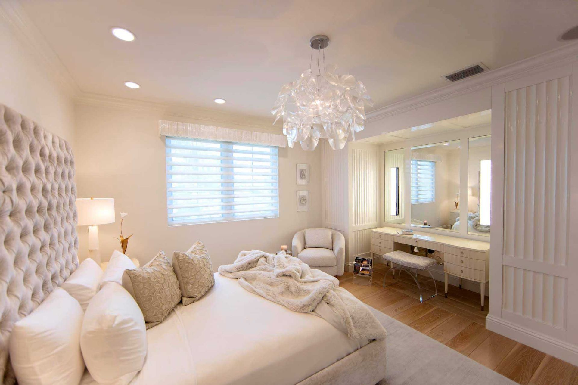 Elegant white bedroom with tufted headboard, vanity, chandelier, and armchair.