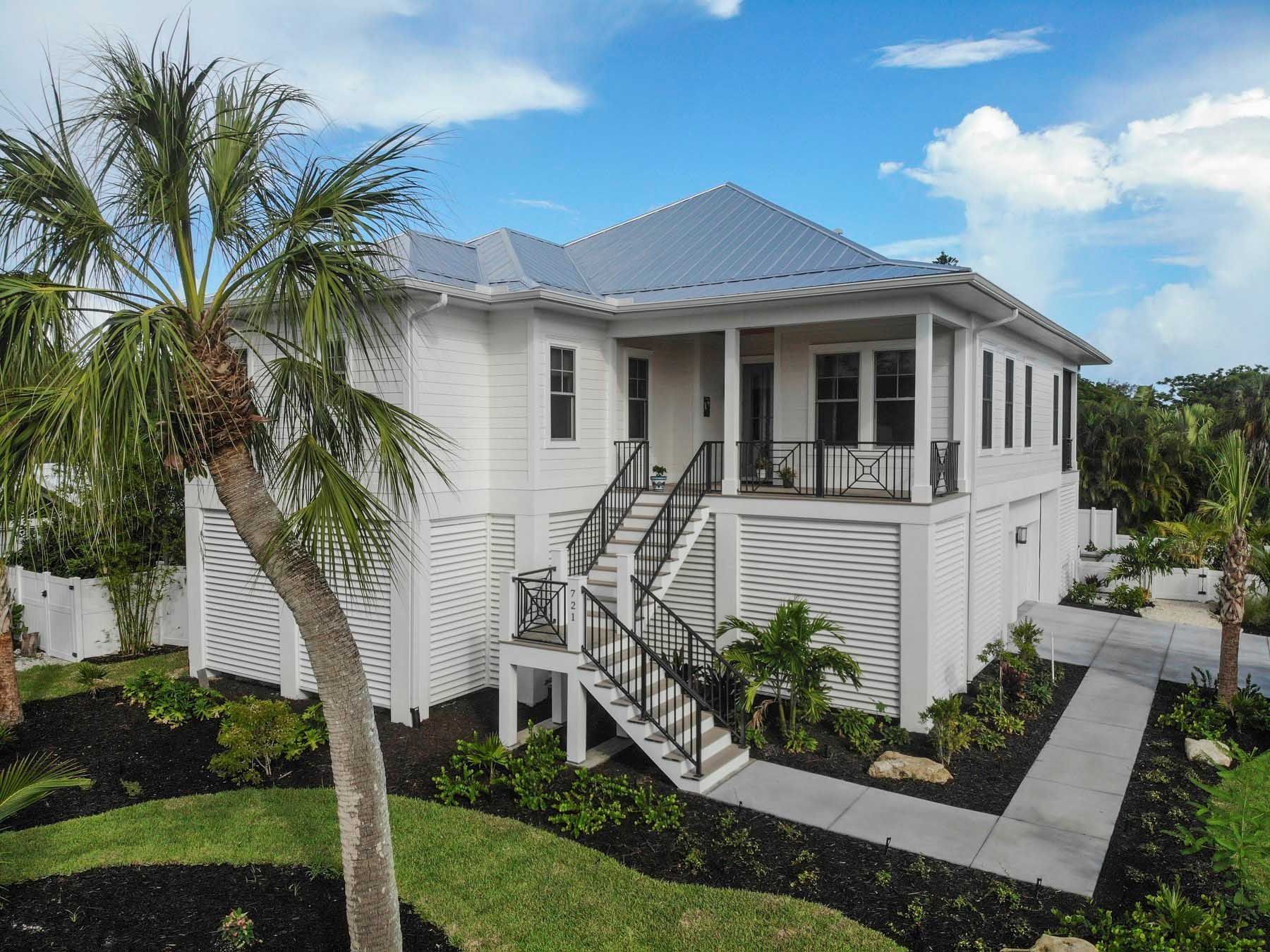 White two-story house with black railings, palm tree, and pathway on a sunny day.