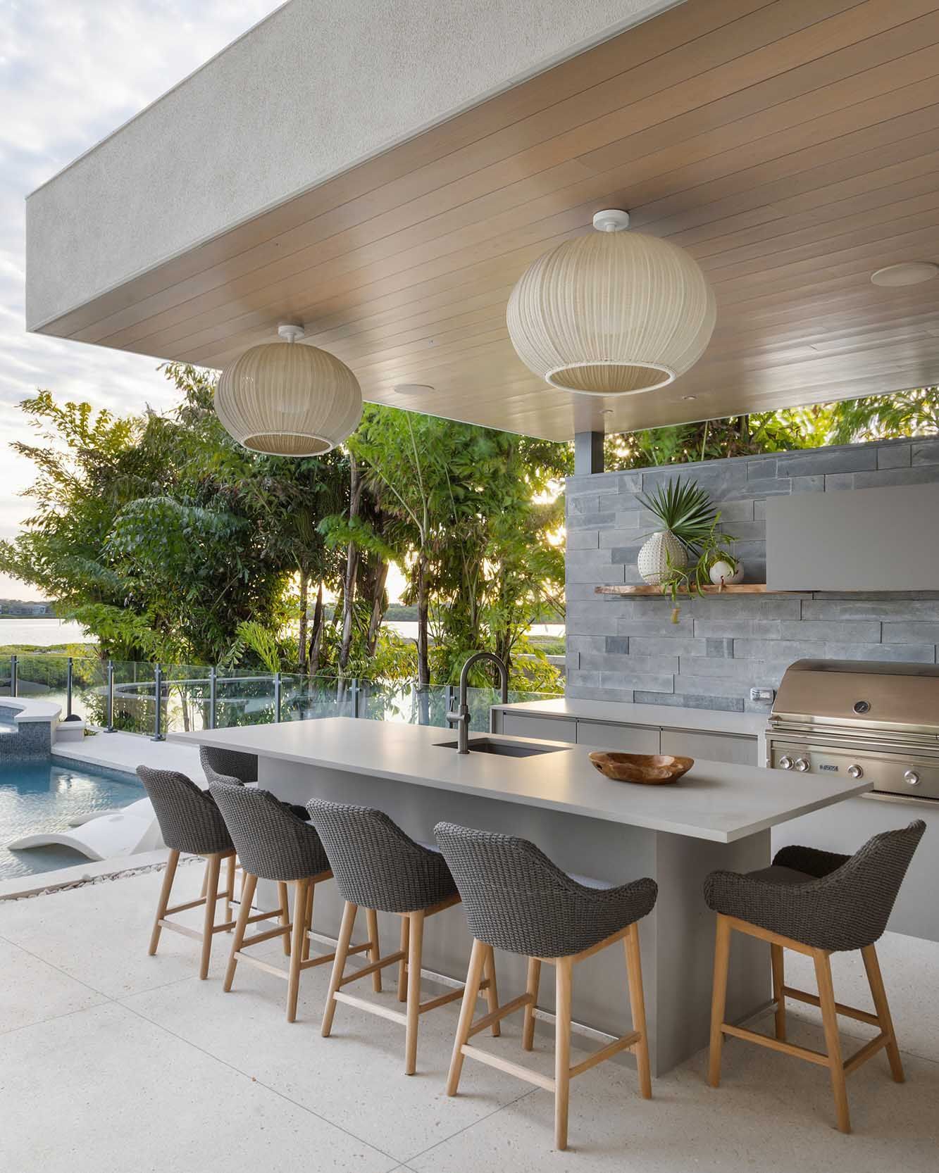 Outdoor kitchen with a bar, stools, and pendant lights overlooking a pool and foliage.
