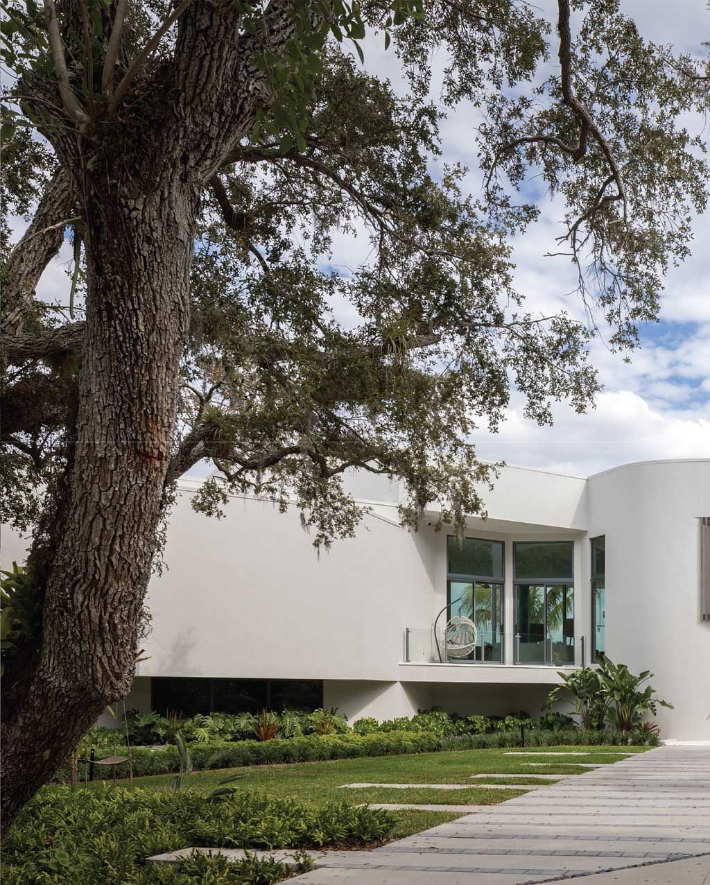 White curved modern house with large tree in front. Lush landscaping, cloudy sky.