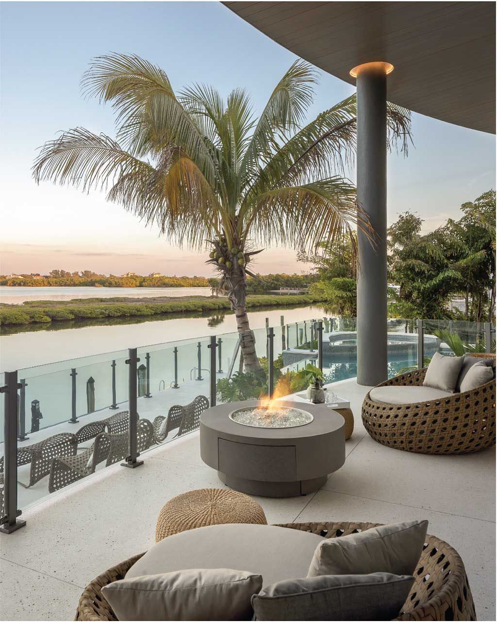 Outdoor patio overlooking water with fire pit, woven seating, palm tree, and glass railing.